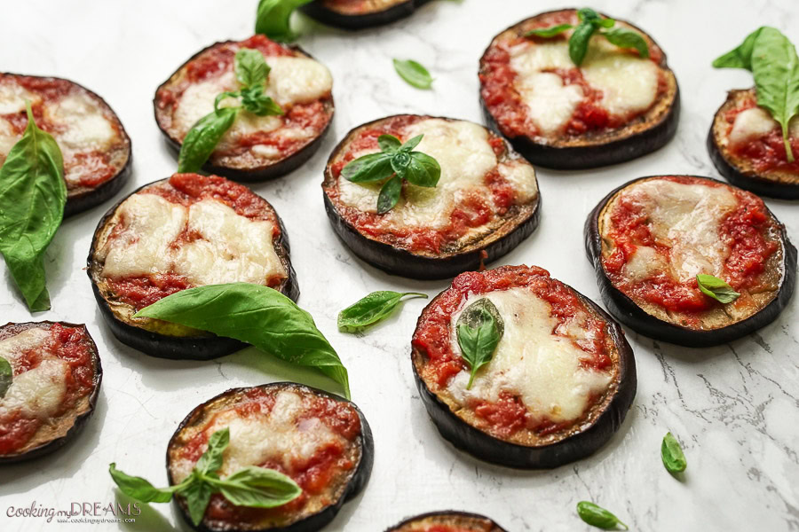 Slices of baked eggplant topped with tomato sauce, melted cheese, and fresh basil leaves are arranged on a white surface.