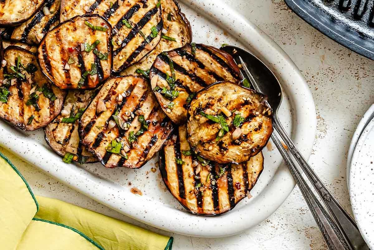 Grilled eggplant slices with visible char marks, garnished with chopped herbs, served on a white oval platter with two metal serving spoons.