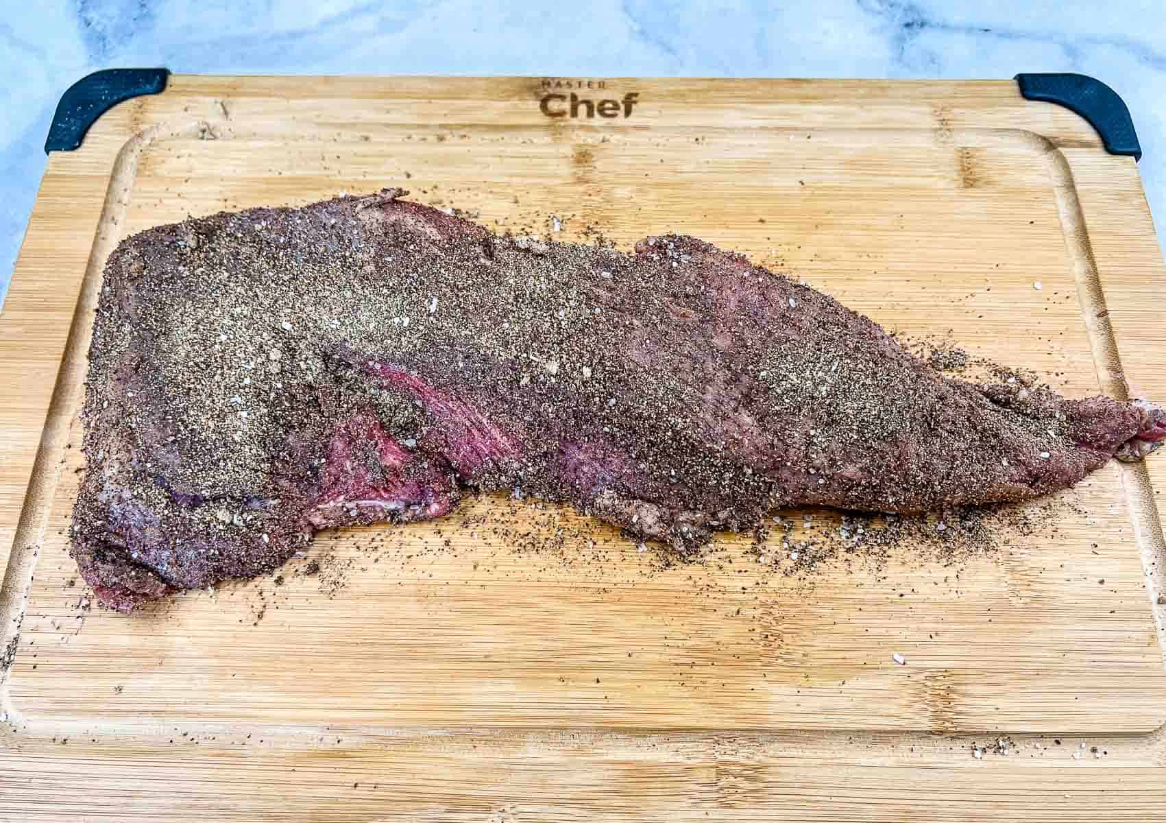 A raw beef tri-tip covered in dry seasoning sits on a wooden cutting board with black corner grips.