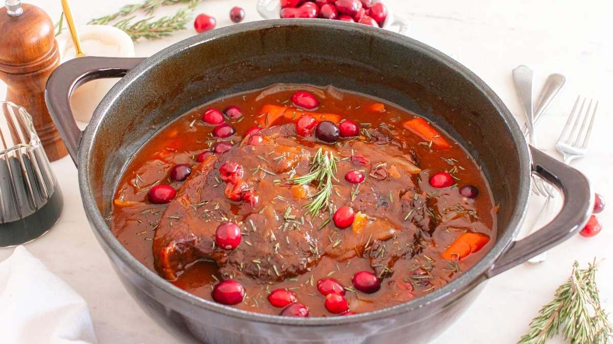 A pot roast with carrots and cranberries in rich brown sauce, garnished with rosemary, sits in a black Dutch oven on a white table.