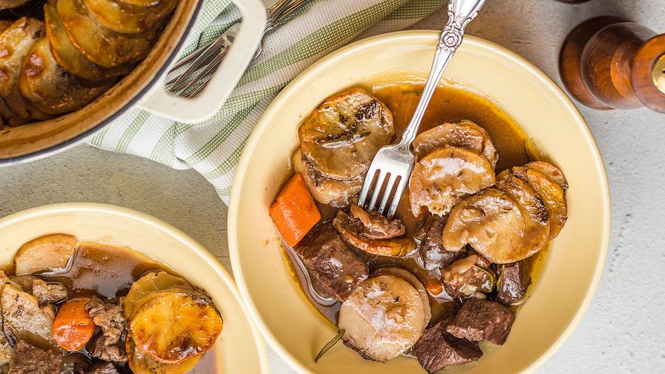 A bowl of beef stew with sliced potatoes, carrots, and sauce, with a fork resting in the dish. Another bowl and a pot with more stew are nearby.