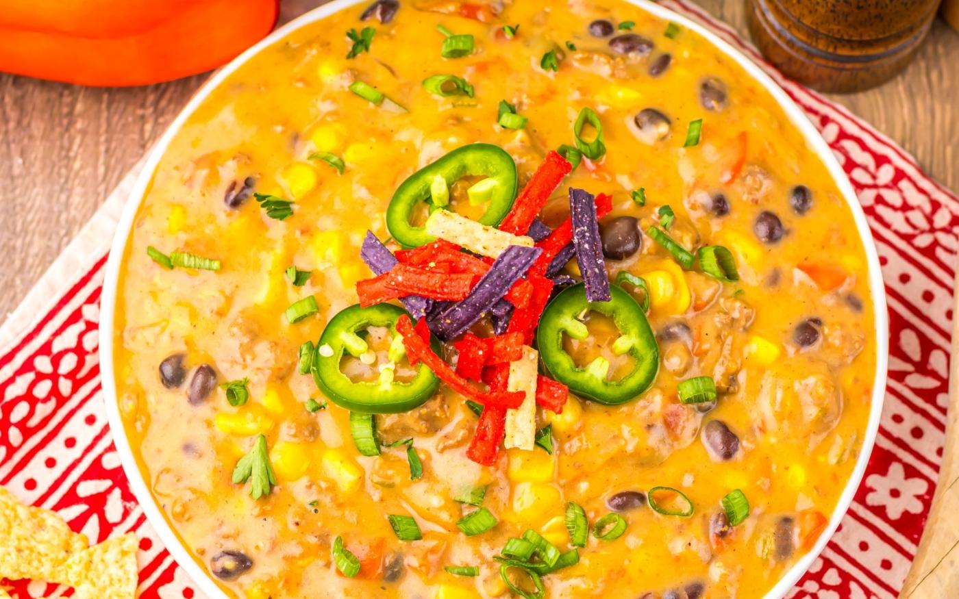 A bowl of creamy Tex-Mex soup with black beans, corn, jalapeño slices, chopped green onions, and colorful tortilla strips, served on a patterned red placemat.