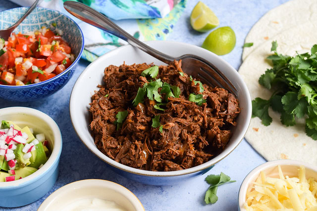 A bowl of shredded meat garnished with cilantro, surrounded by tortillas, chopped vegetables, shredded cheese, salsa, and lime wedges on a blue surface.