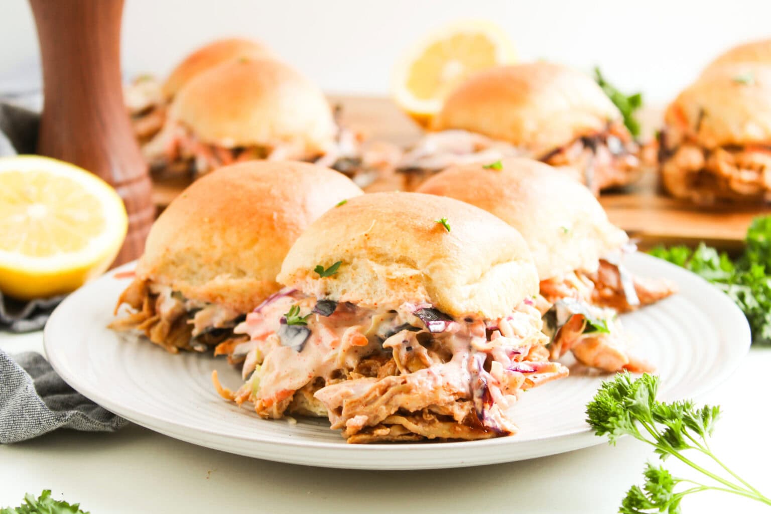 Four pulled pork sliders topped with coleslaw are arranged on a white plate, with more sliders on a wooden board in the background and garnished with parsley and lemon.