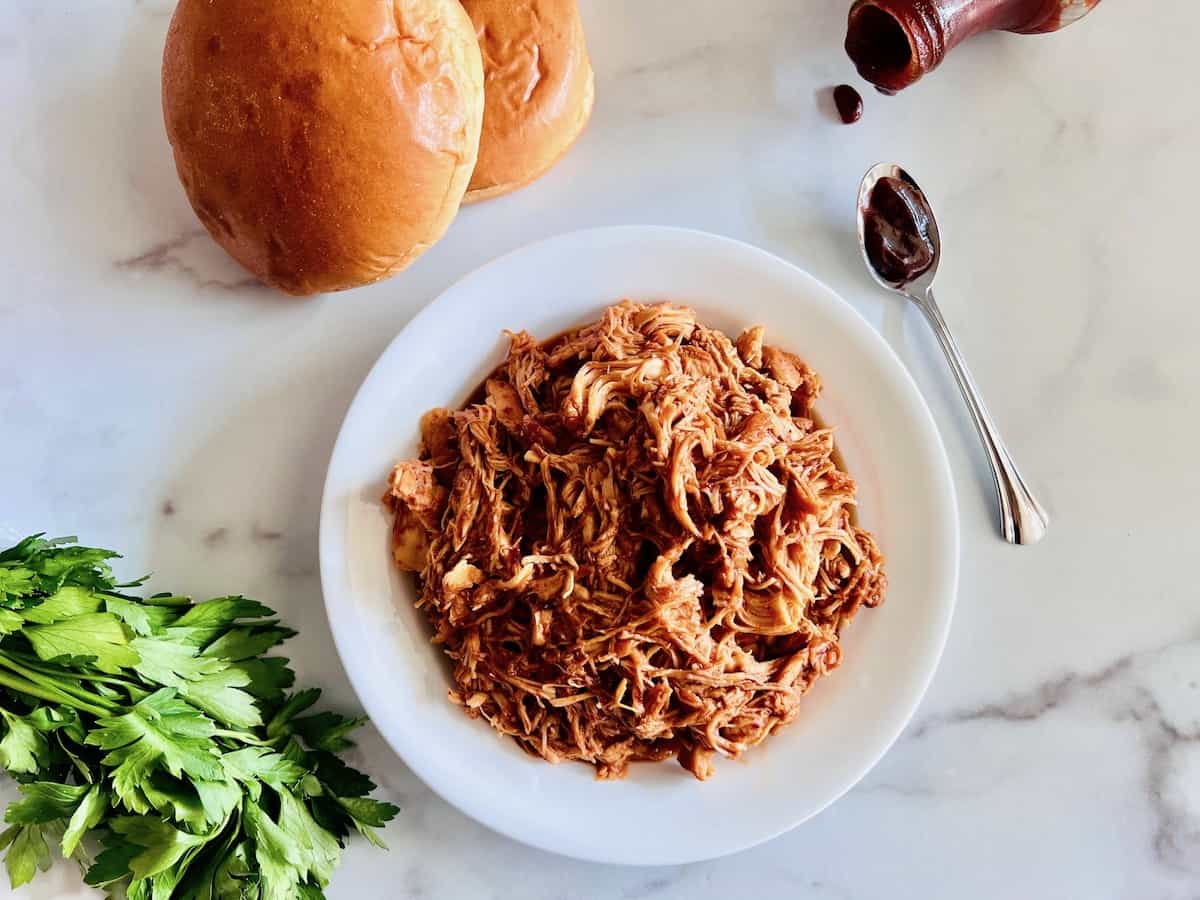 A plate of shredded barbecue chicken sits on a white surface next to two sandwich buns, a bunch of parsley, a spoon, and an open bottle of barbecue sauce.