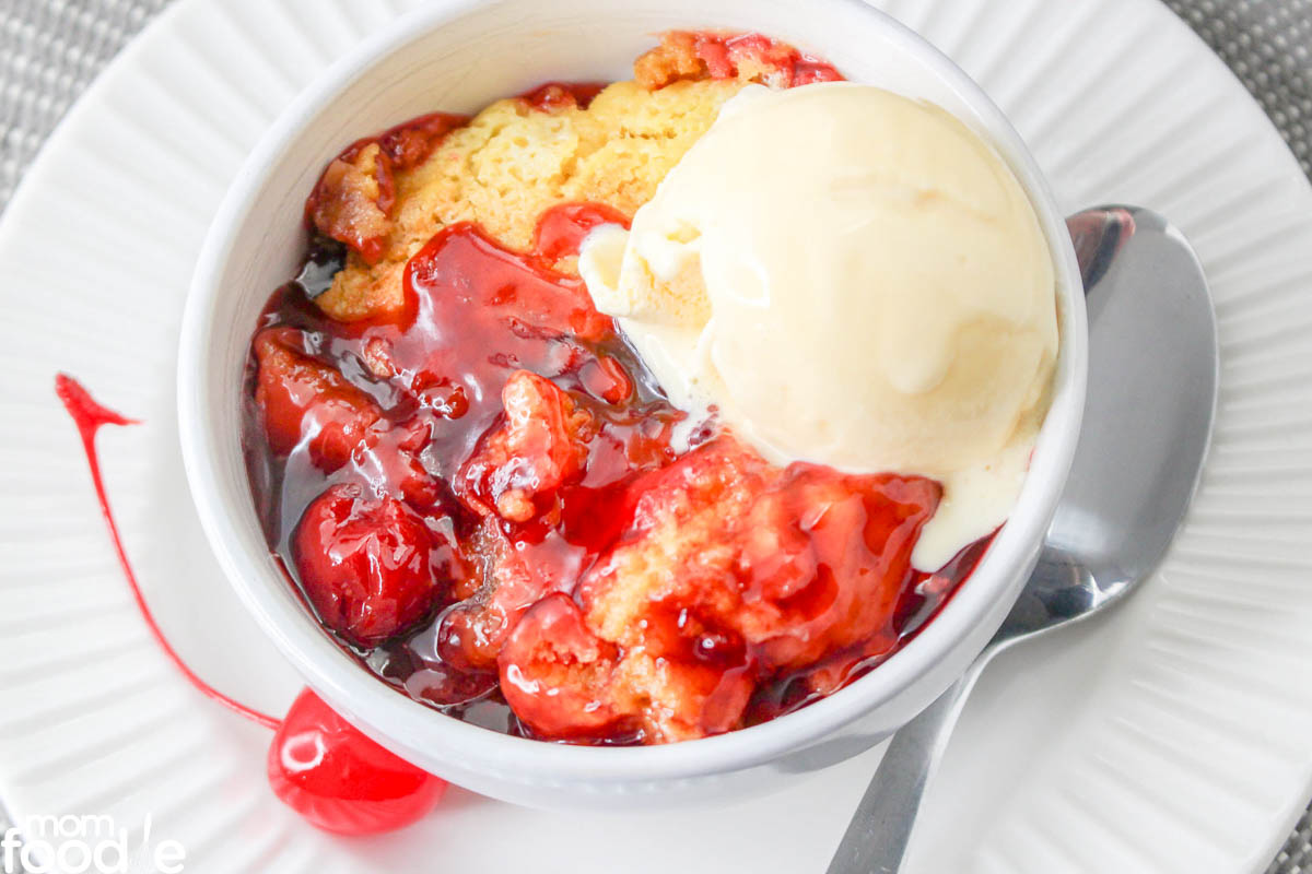 A bowl of cherry cobbler topped with a scoop of vanilla ice cream, served on a white plate with a spoon and a maraschino cherry on the side.
