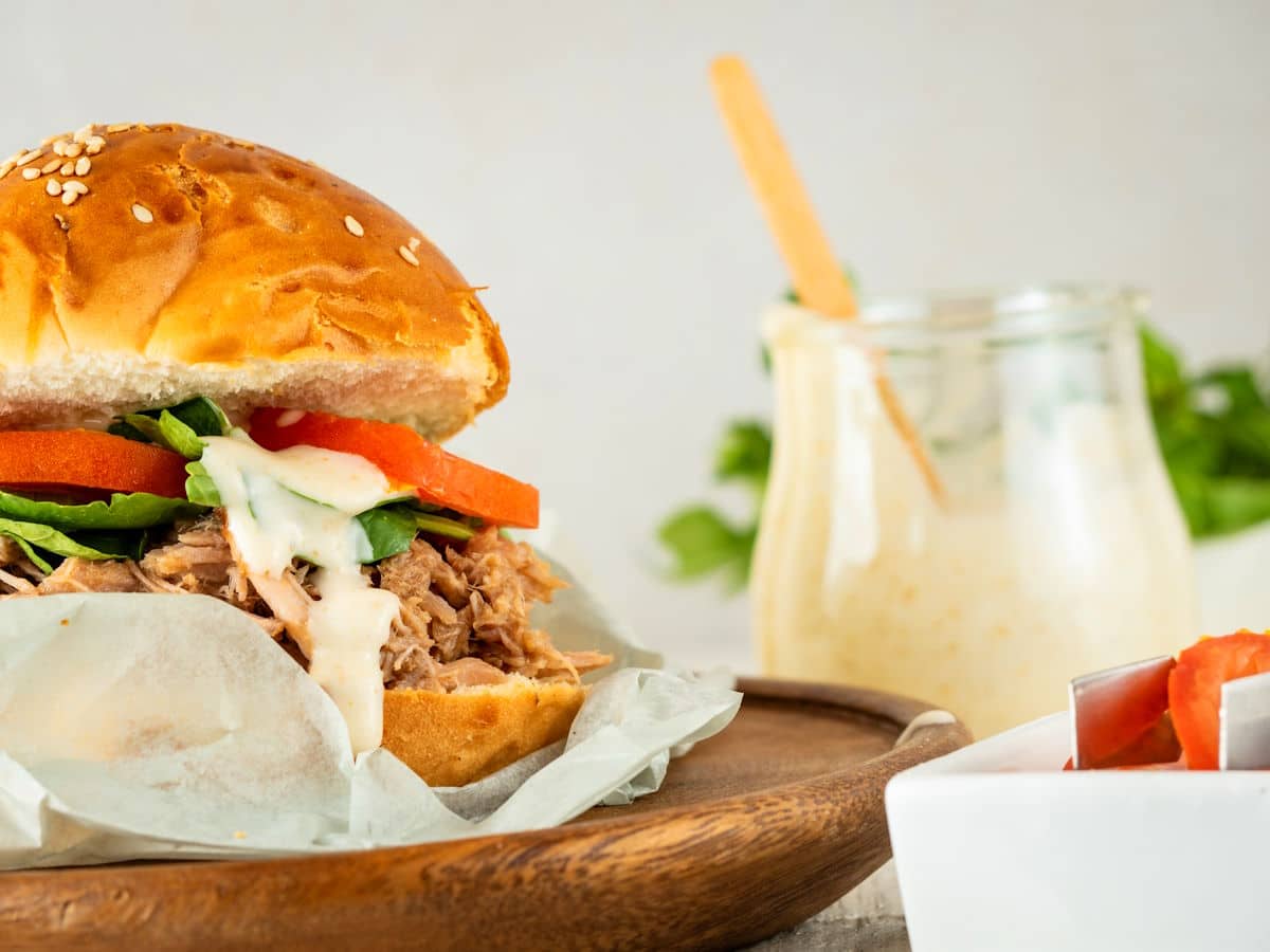 A pulled pork sandwich with lettuce, tomato, and sauce on a bun sits on a wooden plate, with a jar of creamy dressing and a bowl of sliced tomatoes in the background.