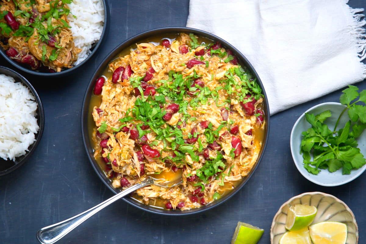 A bowl of shredded chicken stew with red beans, topped with chopped cilantro, next to bowls of white rice, lime wedges, and fresh cilantro on a dark surface.