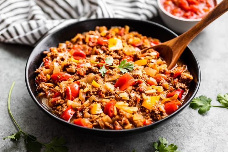 A black bowl filled with a cooked mixture of ground meat, diced tomatoes, onions, and bell peppers, with a wooden spoon and cilantro garnish.