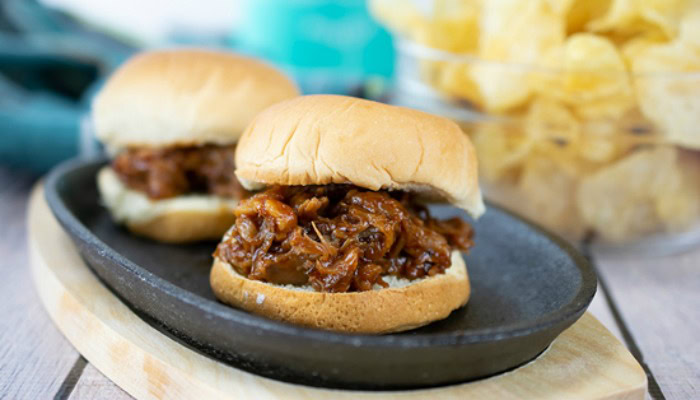 Two pulled pork sliders on buns served on a black plate, with a bowl of potato chips in the background.