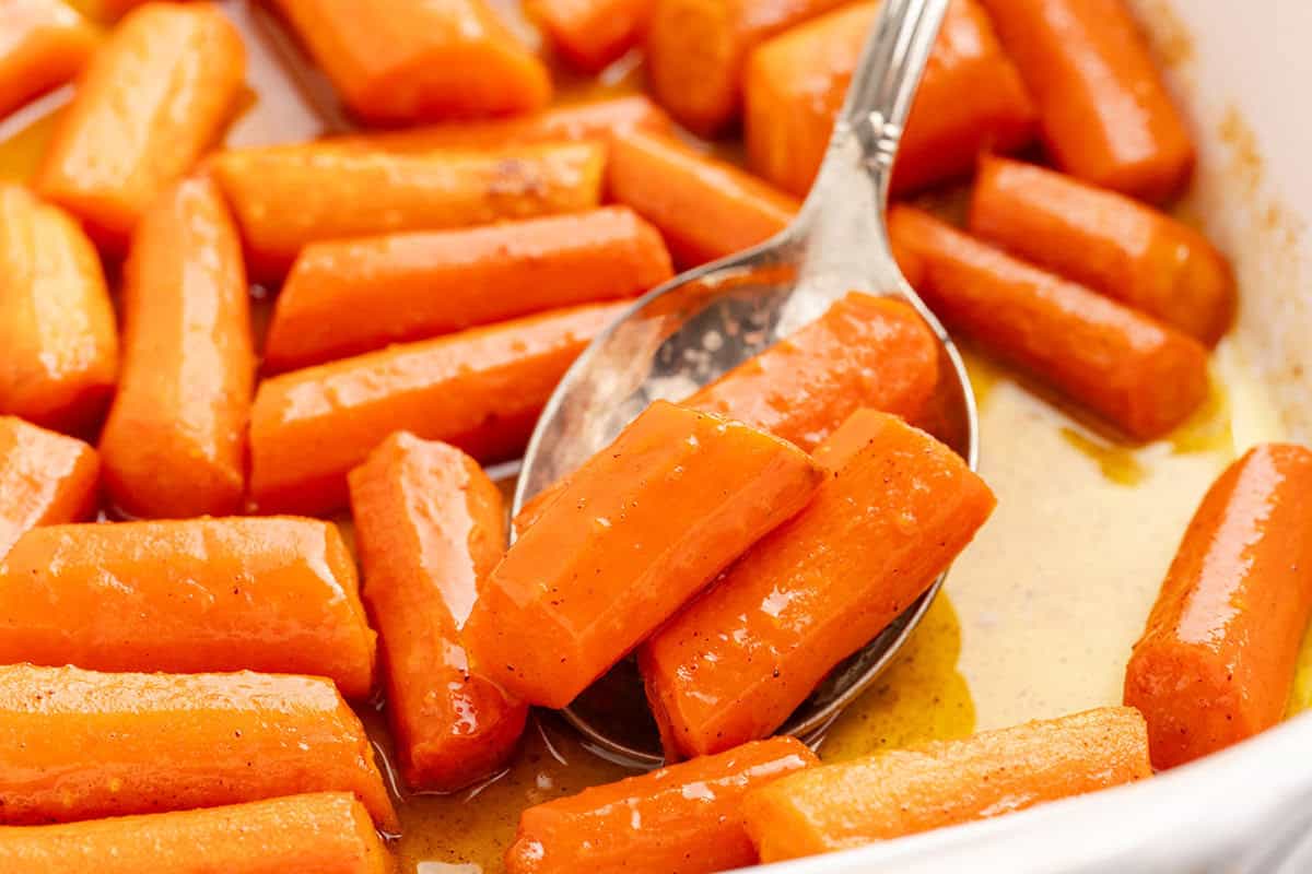 Cooked glazed carrot pieces with a shiny coating are arranged in a dish, with a metal spoon scooping up several pieces.