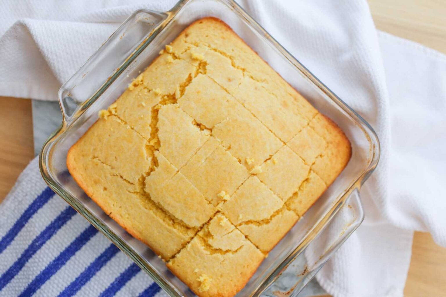 A glass baking dish filled with sliced cornbread rests on a white towel with a blue striped cloth nearby.