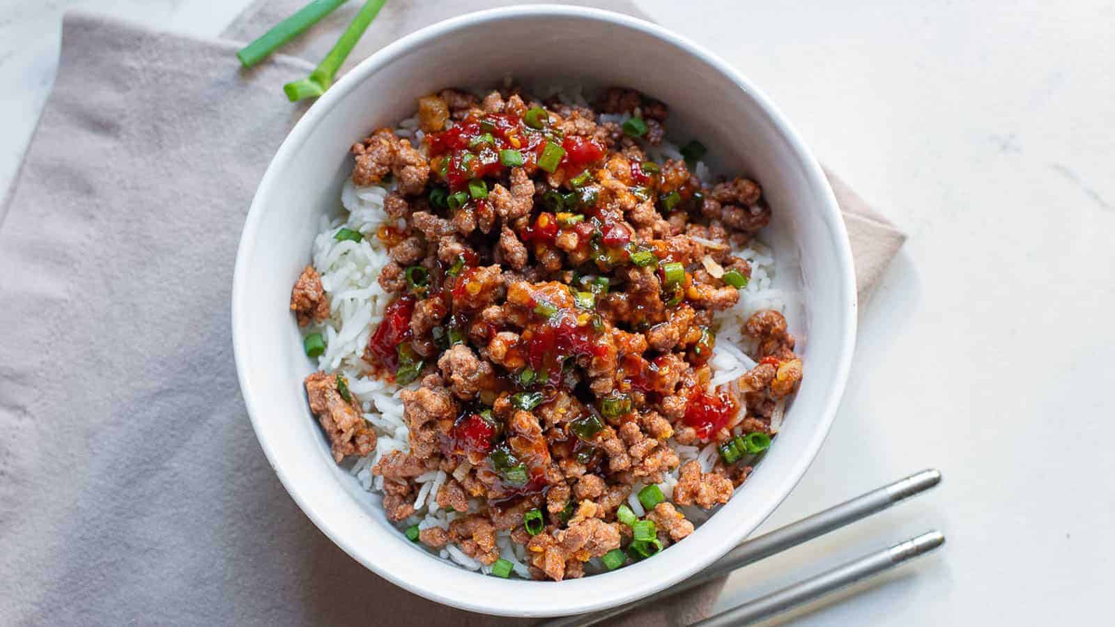 A white bowl filled with steamed rice, topped with ground meat, chopped green onions, and a red sauce, placed on a napkin with chopsticks beside it.
