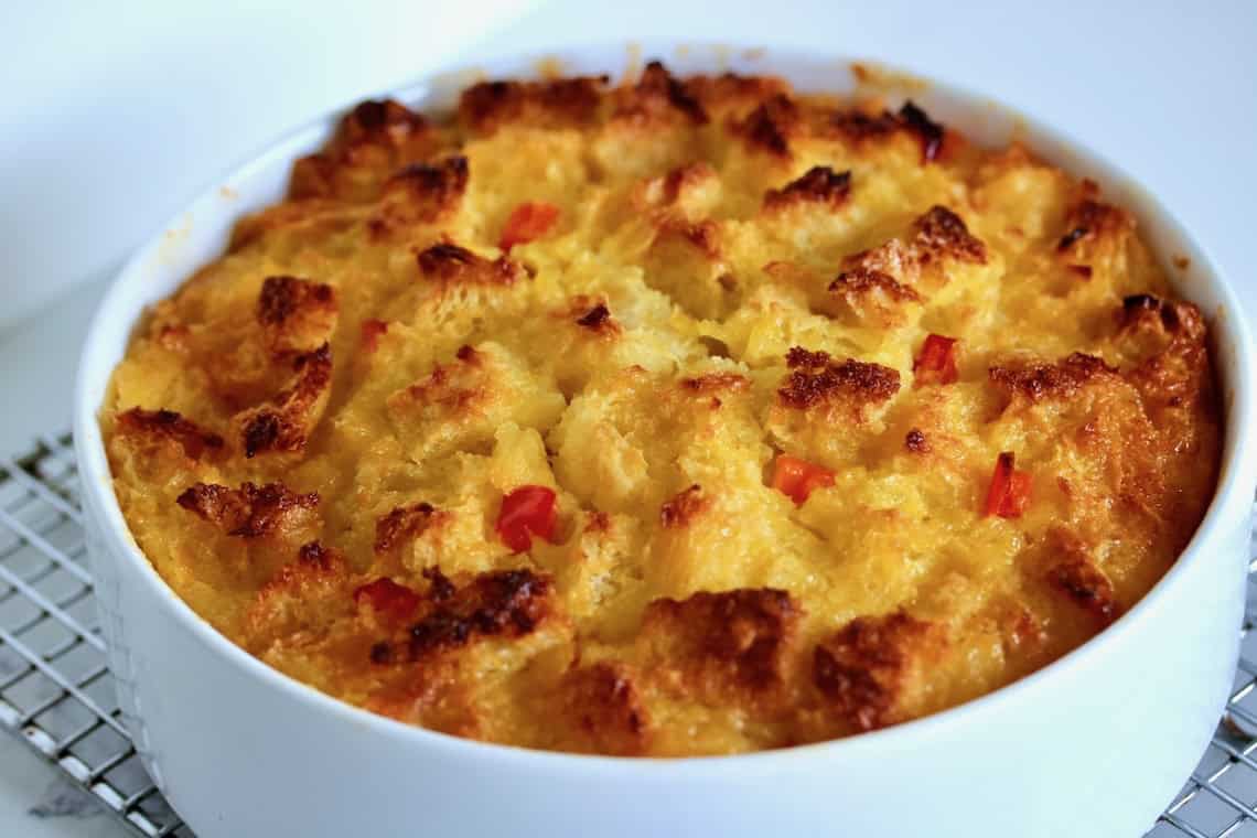 A baked bread pudding with a golden-brown, crispy top and visible pieces of bread and red bell pepper in a round white dish.