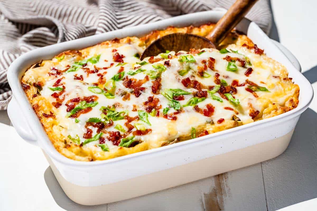 A baked casserole topped with melted cheese, chopped green onions, and bits of bacon, with a serving spoon in the dish.
