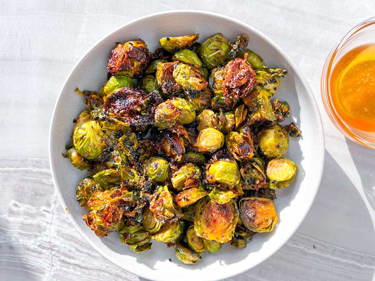 A bowl of roasted Brussels sprouts with crispy, browned edges sits on a light-colored surface next to a small glass bowl containing a golden-colored sauce.