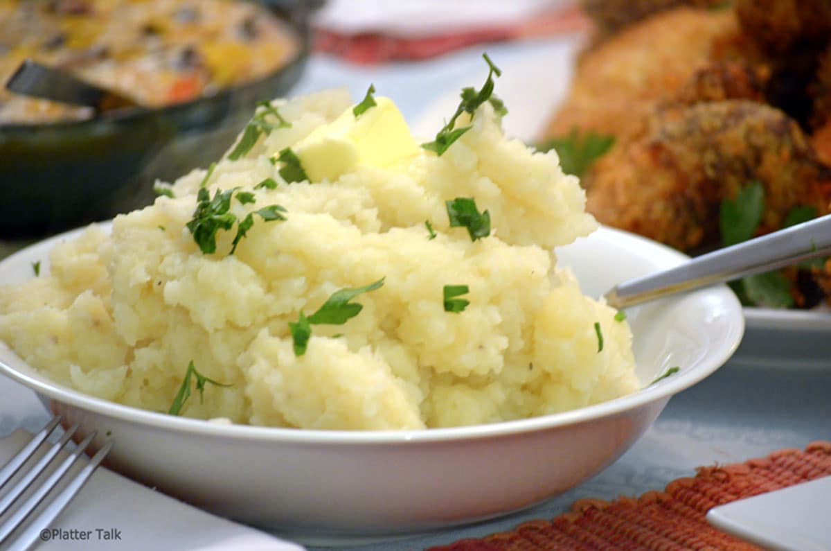 A bowl of mashed potatoes topped with a pat of butter and sprinkled with chopped parsley, with a spoon resting on the side.