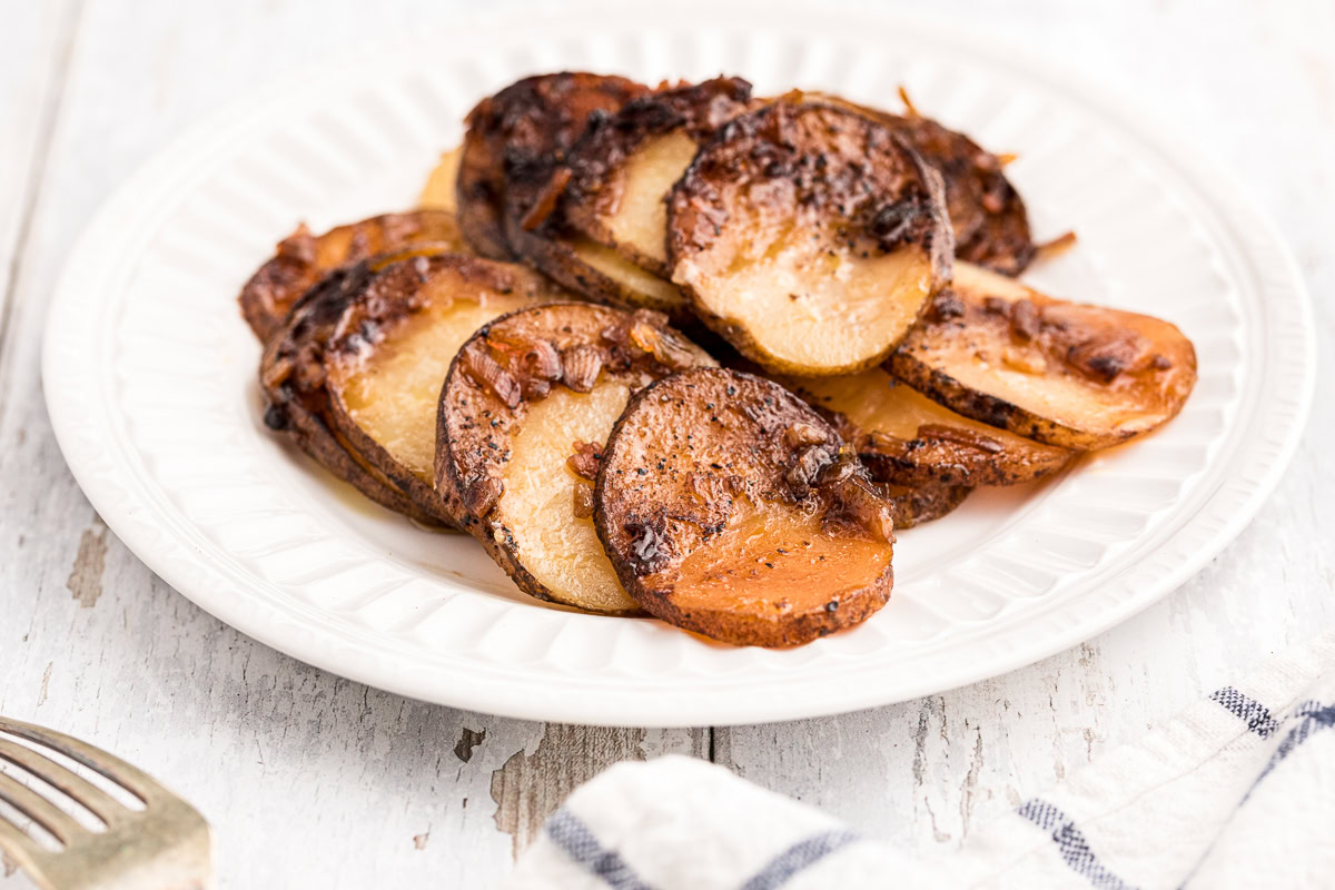 A white plate with sliced, baked potatoes seasoned with a dark brown spice mixture, on a light wooden surface.
