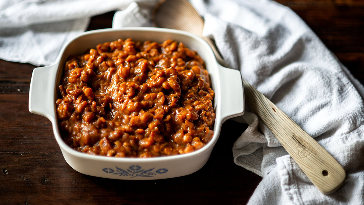 A white casserole dish filled with baked beans sits on a wooden table beside a white towel and a wooden spoon.
