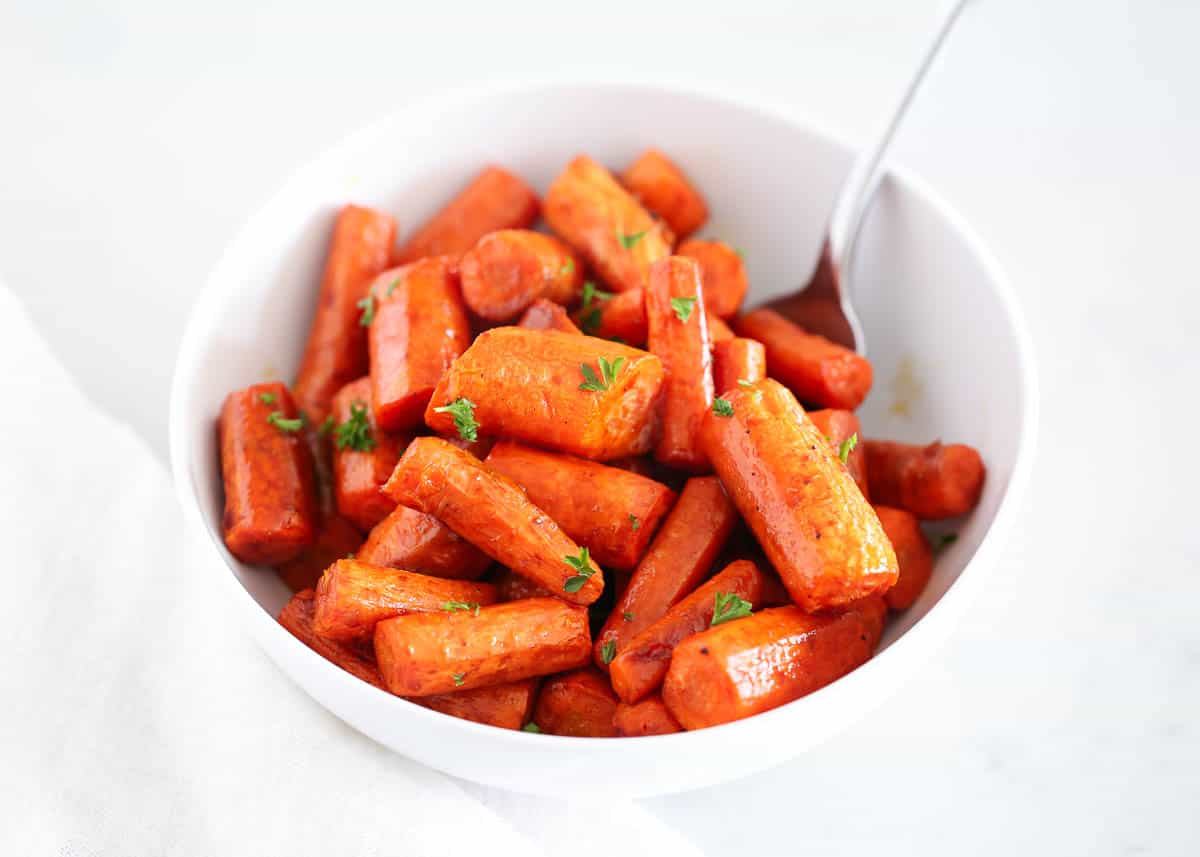 A white bowl filled with roasted carrot pieces garnished with chopped parsley, with a spoon resting inside the bowl.