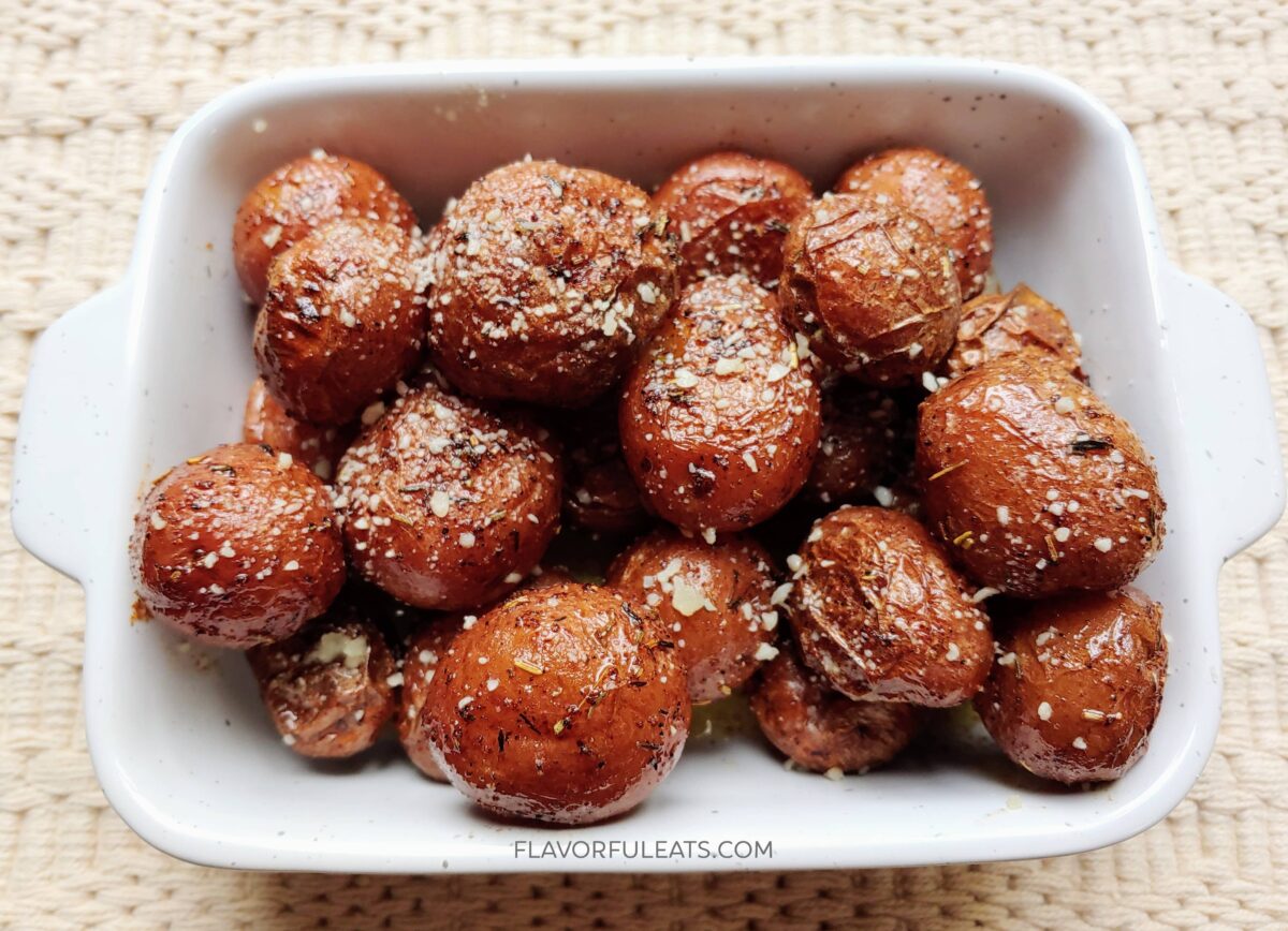 A white baking dish filled with seasoned roasted baby potatoes, sprinkled with grated cheese and herbs, sits on a woven surface.