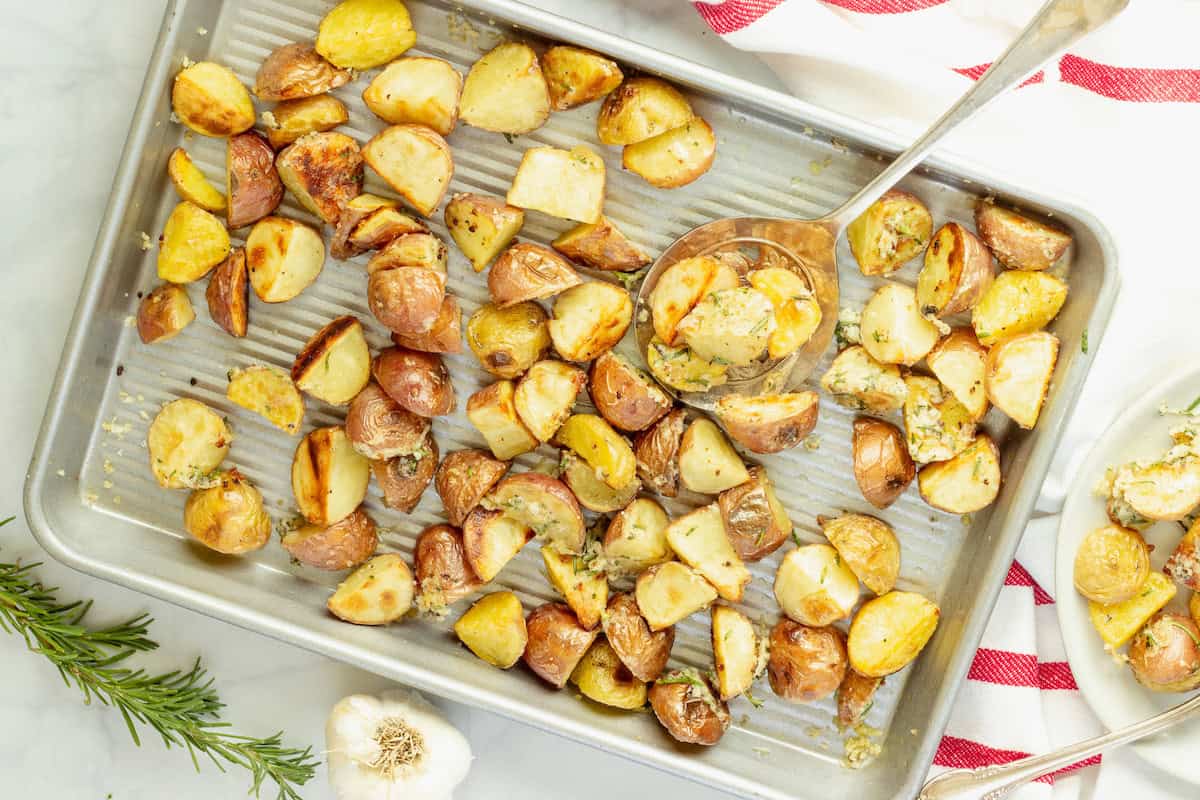 A baking sheet with roasted potato chunks, some served with a large metal spoon; fresh rosemary and a garlic bulb nearby.