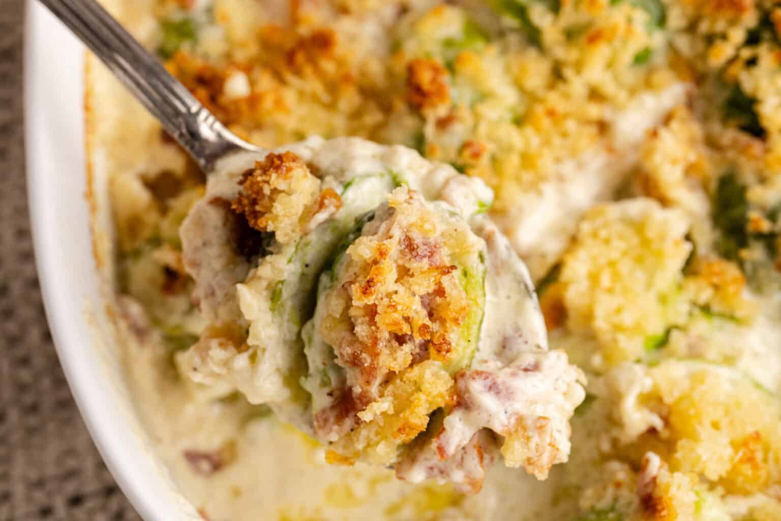A close-up of a spoonful of creamy casserole with a golden breadcrumb topping, being lifted from a baking dish.