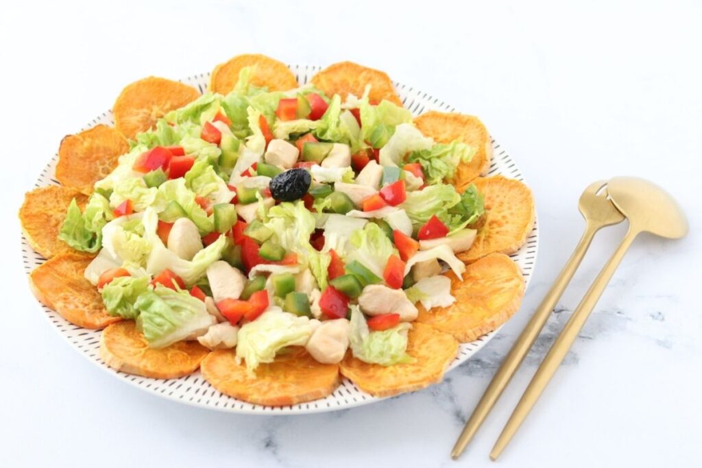 A plate of salad with diced chicken, lettuce, red and green bell peppers, surrounded by sweet potato slices, with gold utensils on the side.