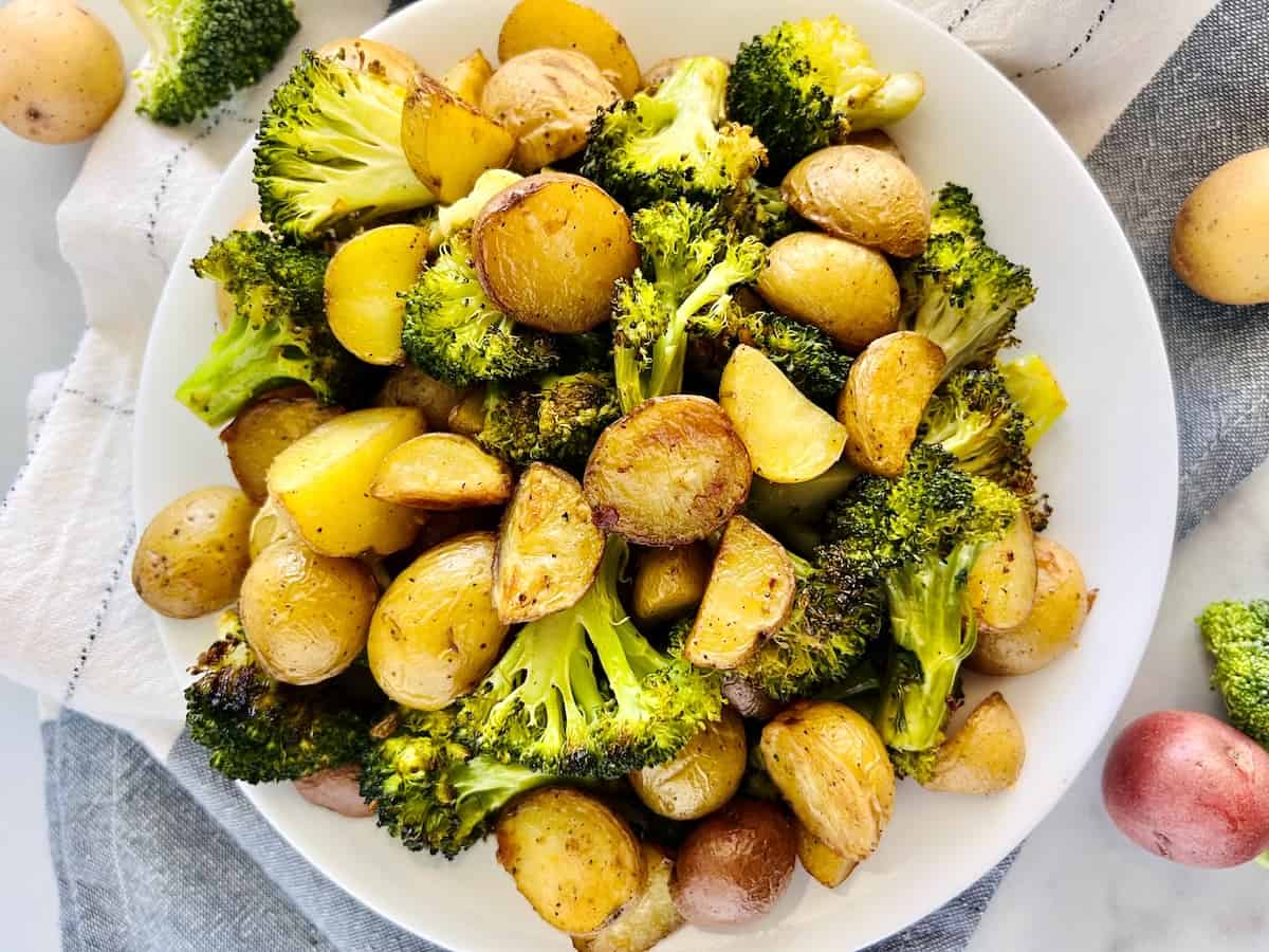 A white plate filled with roasted broccoli florets and halved baby potatoes sits on a light cloth.