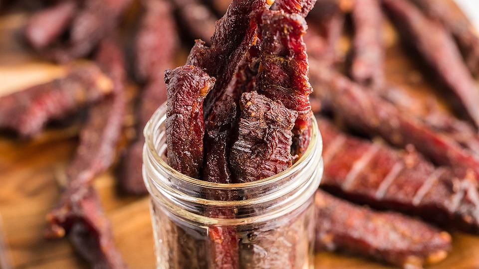 Several pieces of beef jerky are arranged upright in a glass jar, with more jerky pieces scattered on a wooden surface in the background.