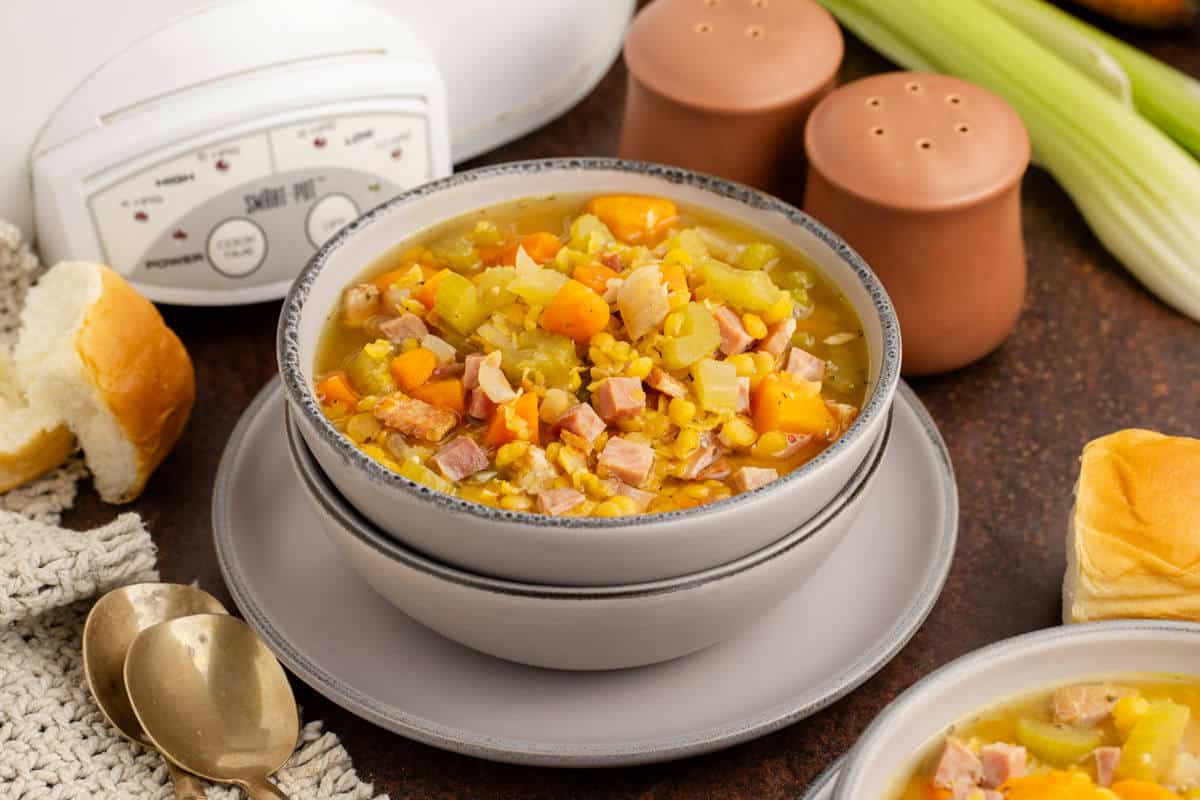 A bowl of split pea soup with diced ham, carrots, and celery sits on stacked plates, next to bread rolls, salt and pepper shakers, spoons, and a slow cooker.