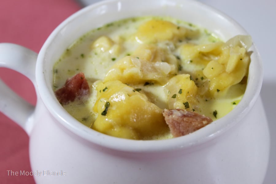 A white bowl filled with creamy soup containing dumplings, pieces of meat, and herbs, with a pink surface in the background.