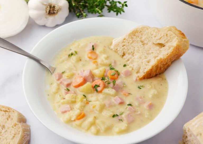 A bowl of creamy soup with diced ham, carrots, and potatoes, garnished with herbs, served with two slices of bread on the side.