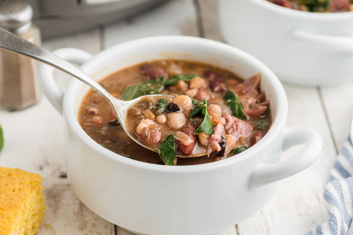 A white bowl filled with bean and vegetable soup, with a spoon lifting a portion. A piece of cornbread and another bowl are nearby on a wooden surface.