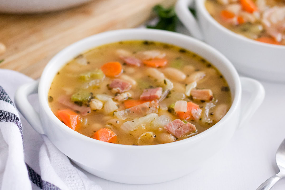 A white bowl filled with ham and bean soup, containing chopped carrots, celery, white beans, and herbs, placed on a white surface.