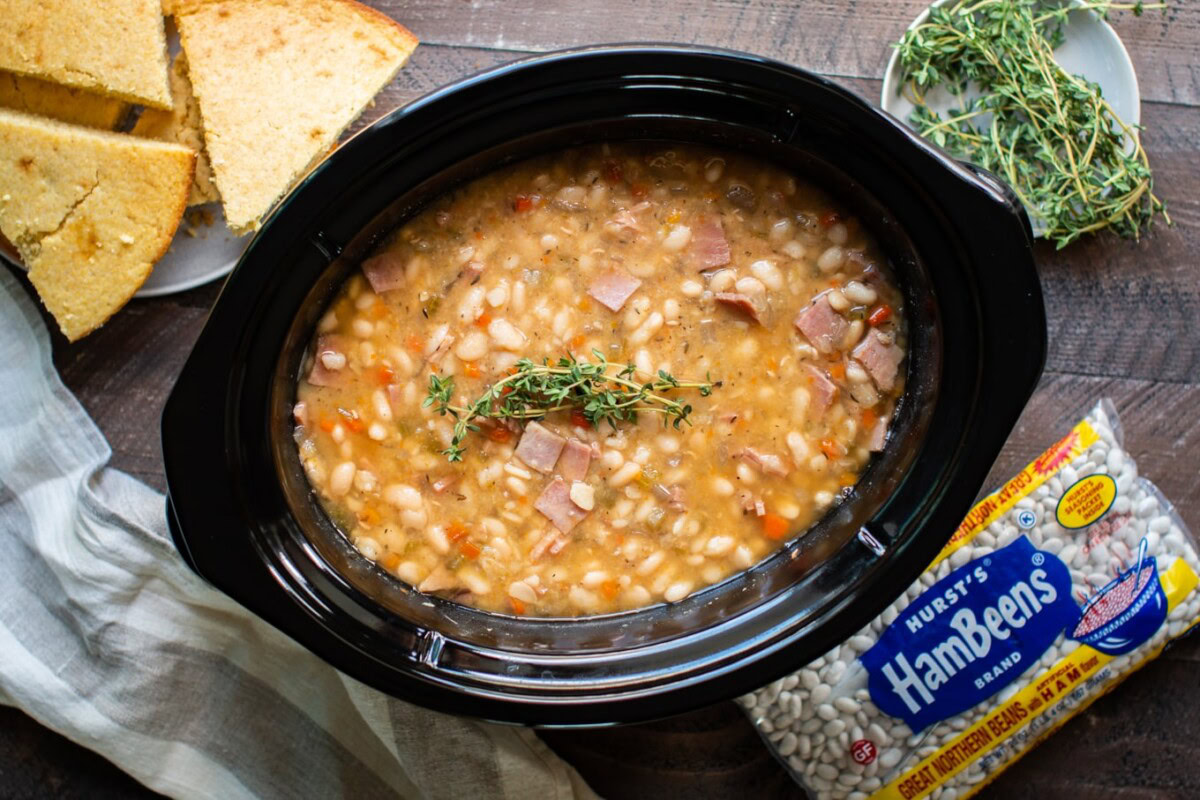 A slow cooker filled with ham and bean soup garnished with fresh thyme, accompanied by cornbread slices and a bag of dried great northern beans on a wooden table.