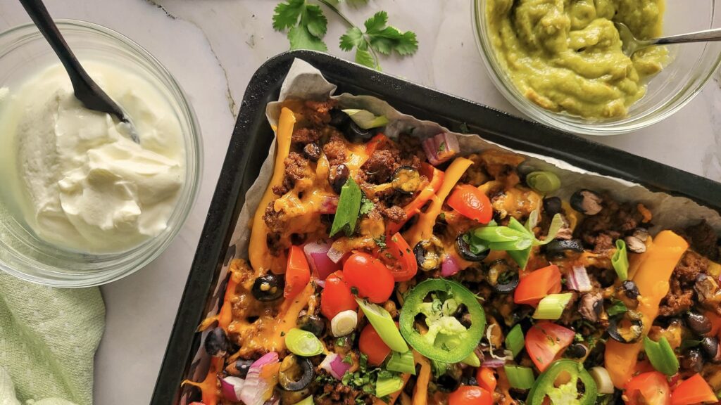 A tray of loaded nachos with ground beef, cheese, tomatoes, olives, onions, and jalapeรฑos, next to bowls of sour cream and guacamole.