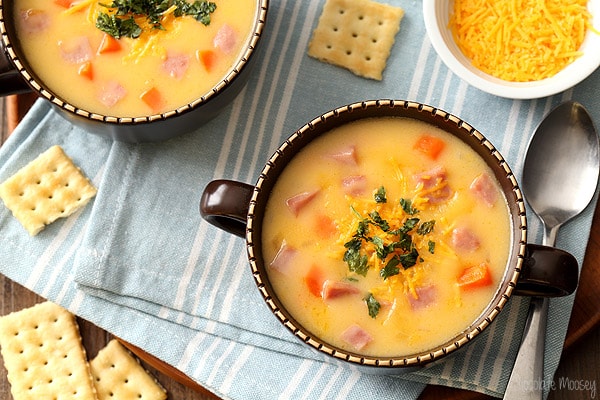 Two bowls of creamy soup with diced ham, carrots, and herbs, served on a striped cloth with saltine crackers and shredded cheese on the side.