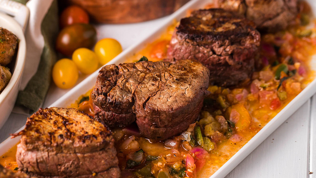Three grilled steaks are served on a rectangular white plate over a bed of chopped tomatoes, onions, and green peppers, with cherry tomatoes in the background.
