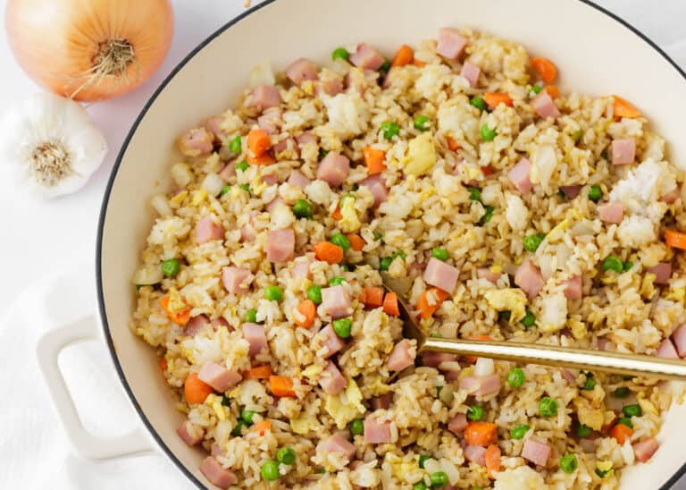 A pan of fried rice with diced ham, peas, carrots, and scrambled egg, with a spoon in the mixture; an onion and a garlic bulb are nearby.