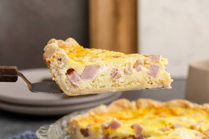A slice of quiche with visible chunks of ham and cheese is held above the rest of the quiche on a metal serving spatula.
