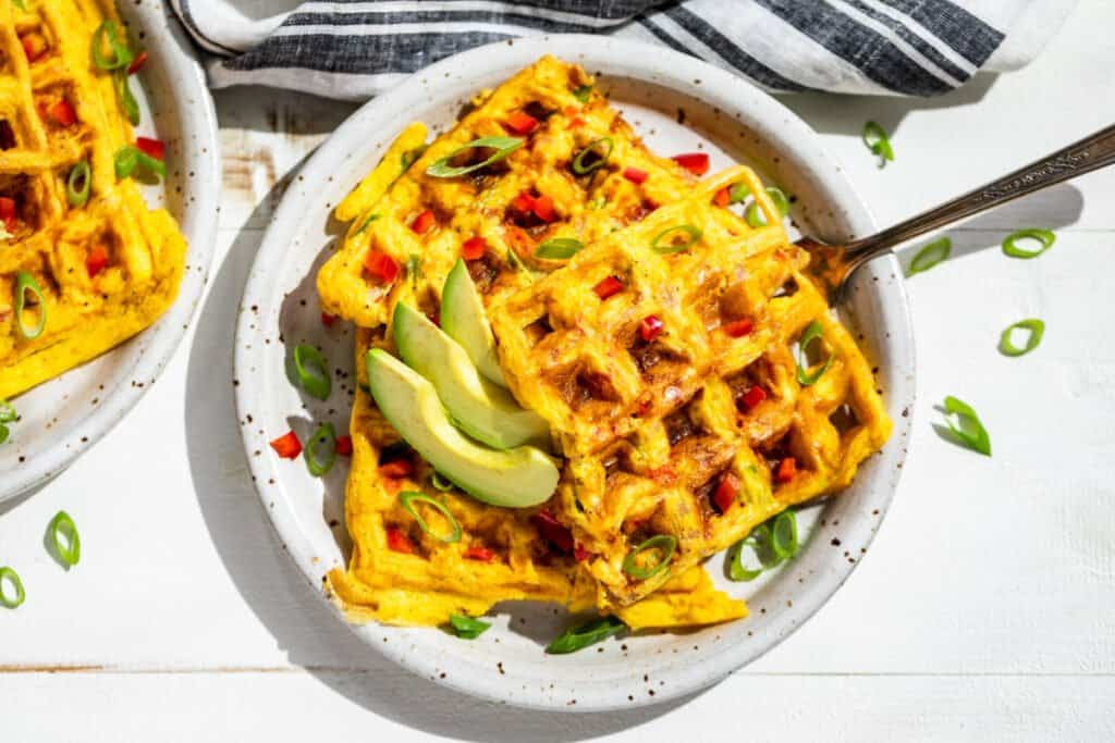 Two savory waffles topped with sliced avocado and chopped green onions are served on a white plate with a fork. A striped napkin is partially visible in the background.
