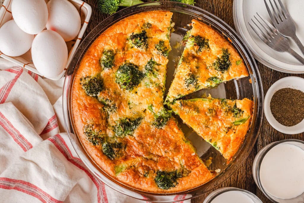 A baked broccoli and cheese quiche in a glass pie dish with two slices cut, surrounded by eggs, seasonings, plates, and utensils on a wooden table.