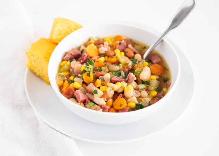 Bowl of ham and bean soup with carrots, corn, and herbs, served with a spoon and two pieces of cornbread on a white plate.