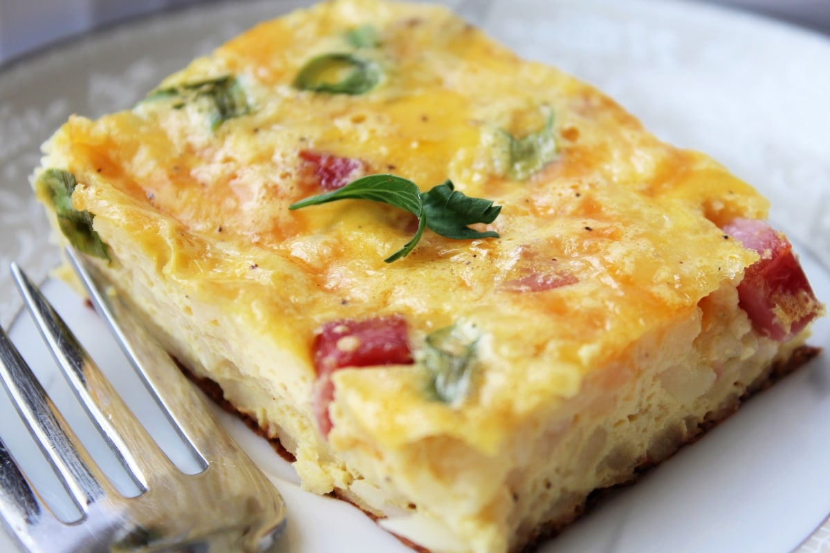 A slice of baked egg casserole with visible diced vegetables and herbs, served on a plate with a fork.