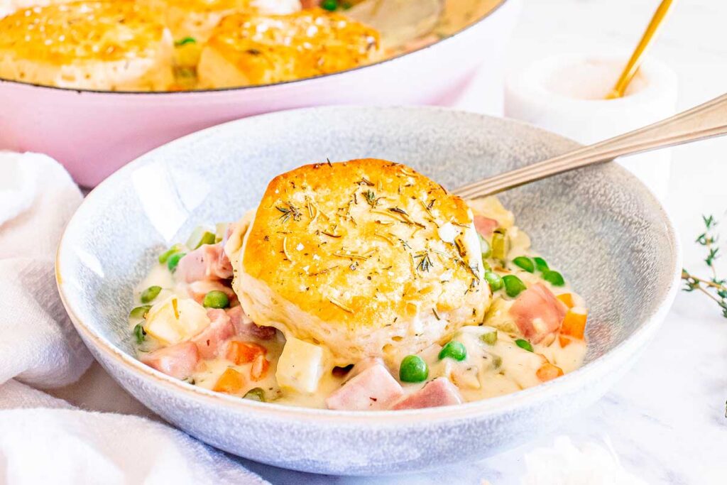 A bowl of creamy chicken pot pie filling with diced ham, peas, carrots, and a large biscuit on top, served with a spoon.