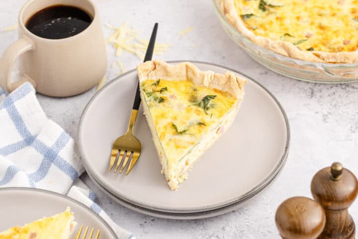 A slice of quiche on a plate with a fork, next to a cup of coffee and a blue-and-white napkin, with a whole quiche and pepper mills in the background.