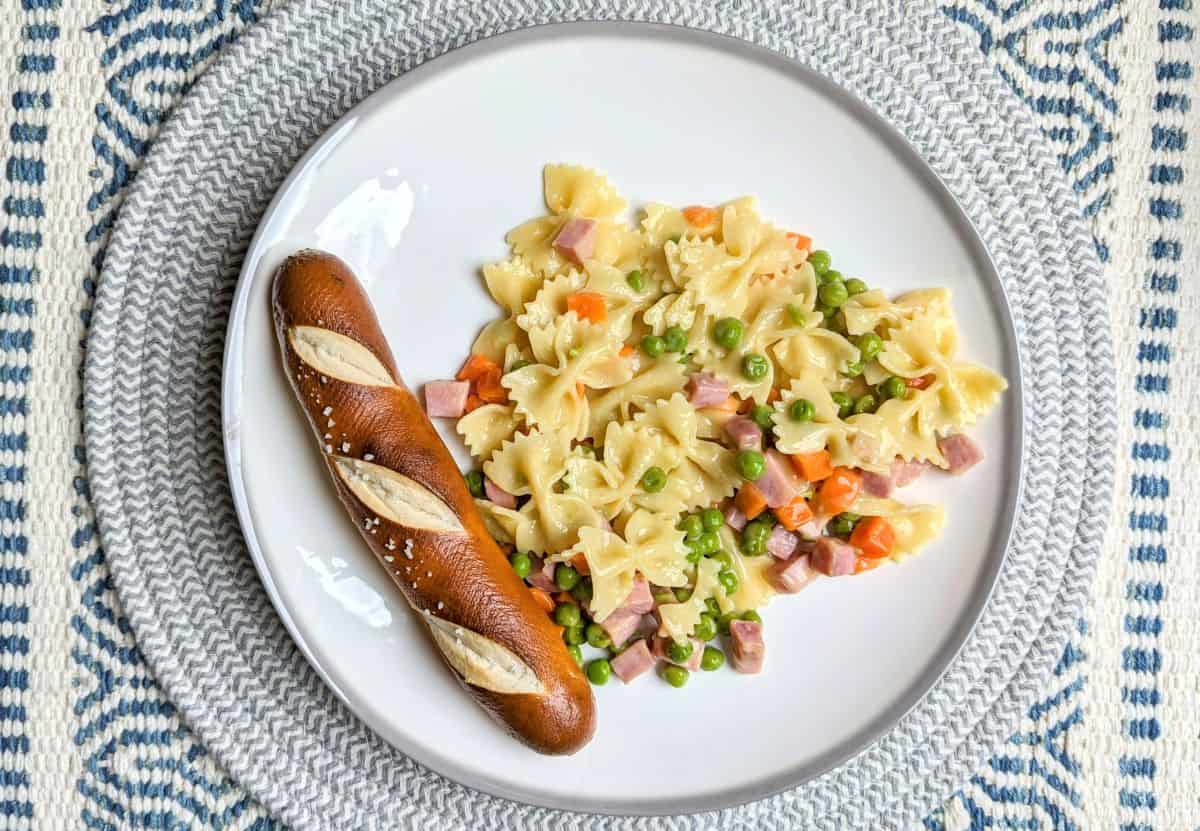 A white plate with bowtie pasta mixed with ham, peas, and carrots, served alongside a salted pretzel roll on a woven placemat.
