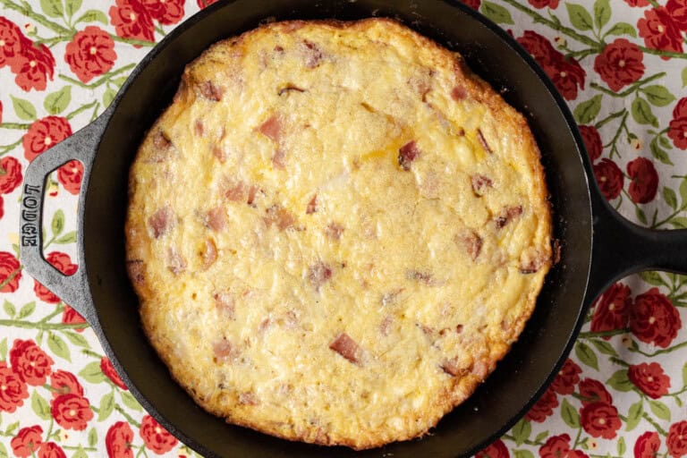 A baked frittata with visible chunks of ham is shown in a black cast iron skillet, placed on a floral tablecloth with red flowers and green leaves.