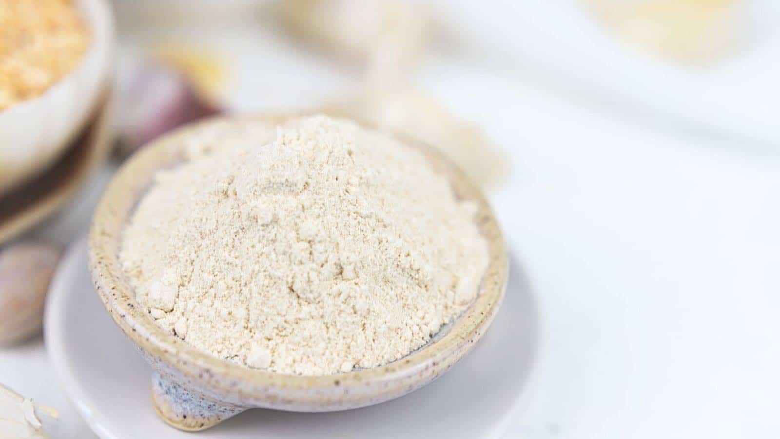 A small ceramic bowl filled with a mound of fine, light-colored powder sits on a white plate against a blurred background.